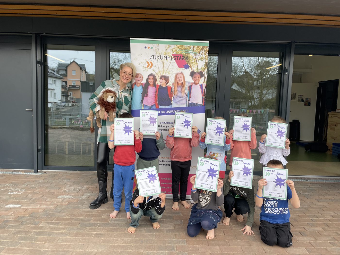 Sarah mit einer Kindergruppe vor dem ZukunftSTARK-Aufsteller, die Kinder halten ihre Urkunden vor sich